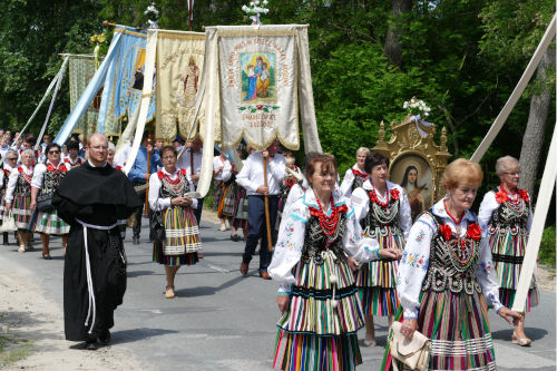 Ludzie idący w procesji Bożego Ciała - kobiety w ludowych strojach, mężczyźni niosący sztandary, obok idący zakonnik.