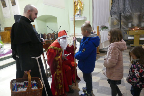 Św. Mikołaj rozdający dzieciom prezenty w kościele. Po jego prawej stronie stoi franciszkanin z koszykiem. 