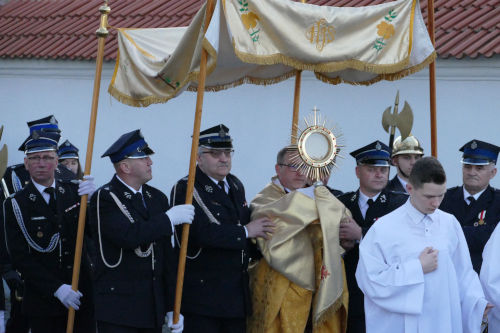 Kapłan niosący Najświętszy Sakrament w asyście ministrantów oraz strażaków.
