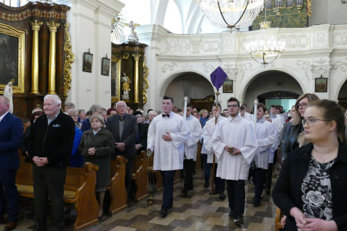 Ministranci wchodzący do kościoła ze świecami oraz krzyżem zasłoniętym puprurową narzutą.