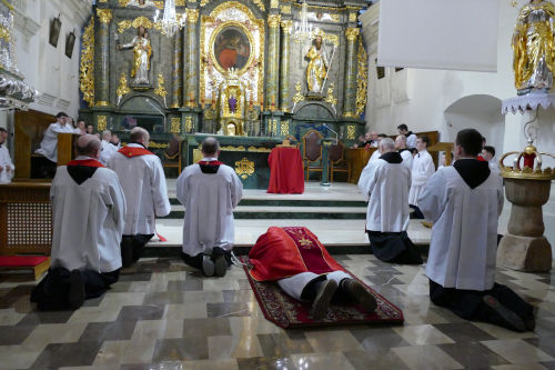 Kapłan leżący na dywanie przed ołtarzem. Po bokach ministranci oraz pozostali kapłani.