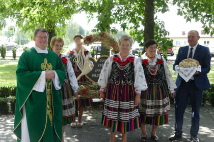 Proboszcz Mariusz Kapczyński stojący z kobietami w strojach ludowych wraz z dożynkowym wieńcem. Po prawej stronie stoi mężczyzna w garniturze trzymający chleb.