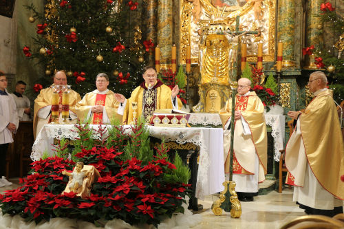 Pięciu Księży w złotych ornatach celebrujących Mszę Świętą przy ołtarzu. Przed Stołem Eucharystyczym znajduje się figurka Jezusa umiejscowiona w czerwonych kwiatach - Gwiazdach betlejemskich. Po lewo w tle choinka oraz ministranci.