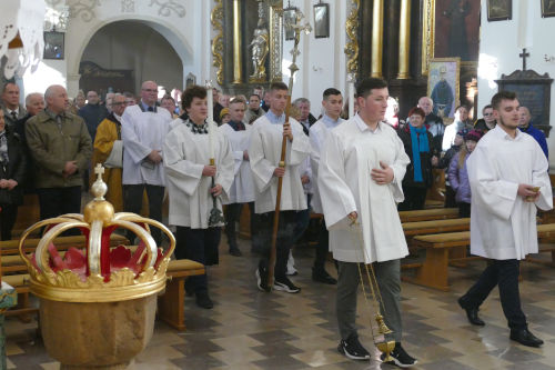 Ministranci wchodzący do kościoła z kadzidłem i Krzyżem. Po lewej stronie na pierwszym planie chrzcielnica. W tle w ławkach stoją wierni.